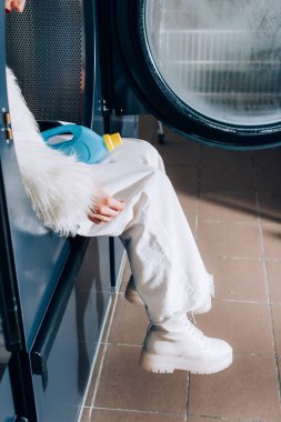 Kırpılmış, elinde deterjanla çamaşır makinesinde oturan kadın manzarası. 