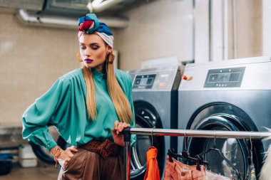Çamaşırhanedeki çamaşır makinelerinin yanında cebinde duran türbanlı şık bir kadın. 