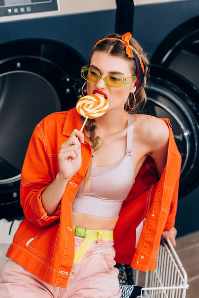 trendy young woman in sunglasses biting lollipop near cart and washing machines in laundromat