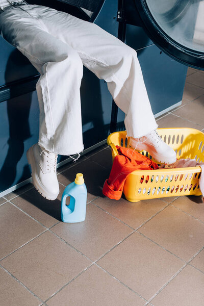 обрезанный вид женщины в белых брюках, сидящей в общественной стиральной машине рядом с корзиной с прачечной и бутылкой моющего средства