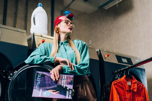 low angle view of trendy woman in glasses and turban holding magazine near washing machines in public laundromat