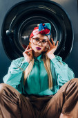 junge Frau mit Brille und Turban blickt in die Kamera neben der Waschmaschine