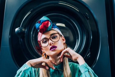 Çamaşır makinesinin yanındaki kameraya bakan gözlüklü ve türbanlı şık genç bir kadın.