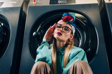 trendy young woman in glasses and turban looking at camera near washing machines clipart