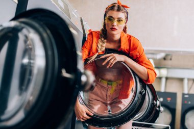 stylish woman in headband and sunglasses near washing machines in modern laundromat with blurred foreground clipart