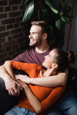 joyful couple embracing while sitting on sofa and looking away clipart