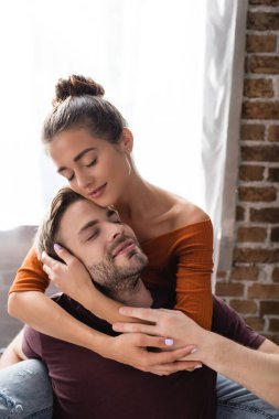 tender woman with closed eyes hugging happy man while sitting at home clipart