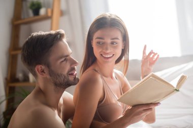 Gülümseyen kadın kitabı tutuyor ve parmağıyla tişörtsüz erkek arkadaşını işaret ediyor.