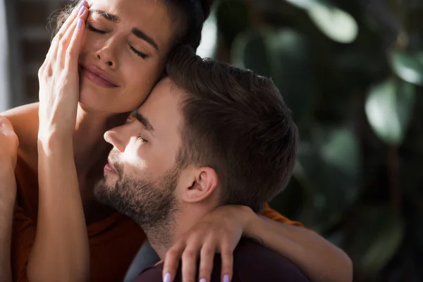 crying woman touching face while embracing beloved man at home