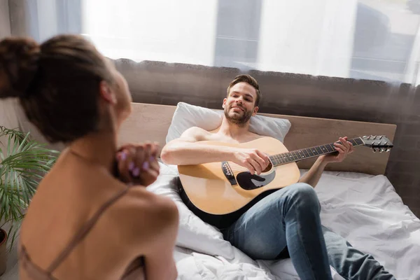 smiling man lying in bed and playing guitar while girlfriend dancing on blurred foreground