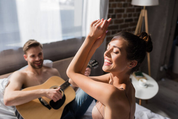 excited woman in bra dancing with closed eyes near boyfriend playing guitar on blurred background