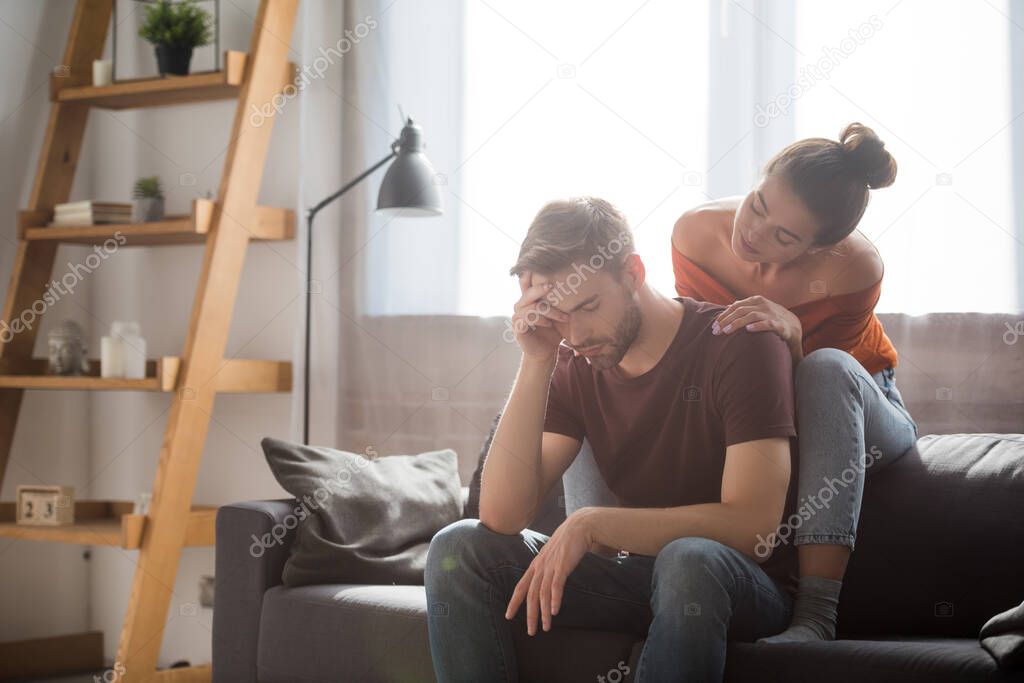 tender woman touching shoulder of worried man sitting on sofa with bowed head and closed eyes