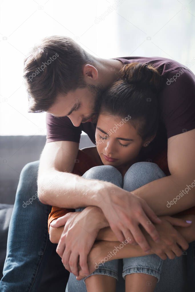 young man embracing and calming beloved woman sitting on sofa with closed eyes