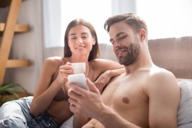 Üstsüz bir adam akıllı telefondan konuşuyor gülümseyen bir kız arkadaşın yanında bulanık arka planda bir fincan kahve tutuyor.