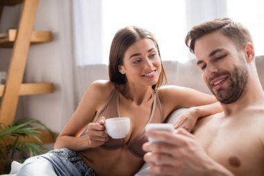 Sütyen giymiş bir kadın, gömleksiz erkek arkadaşına bakarken ön planda bulanık bir şekilde akıllı telefondan sohbet ediyor.