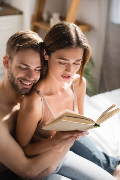 happy man with closed eyes embracing sexy girlfriend reading book in bedroom