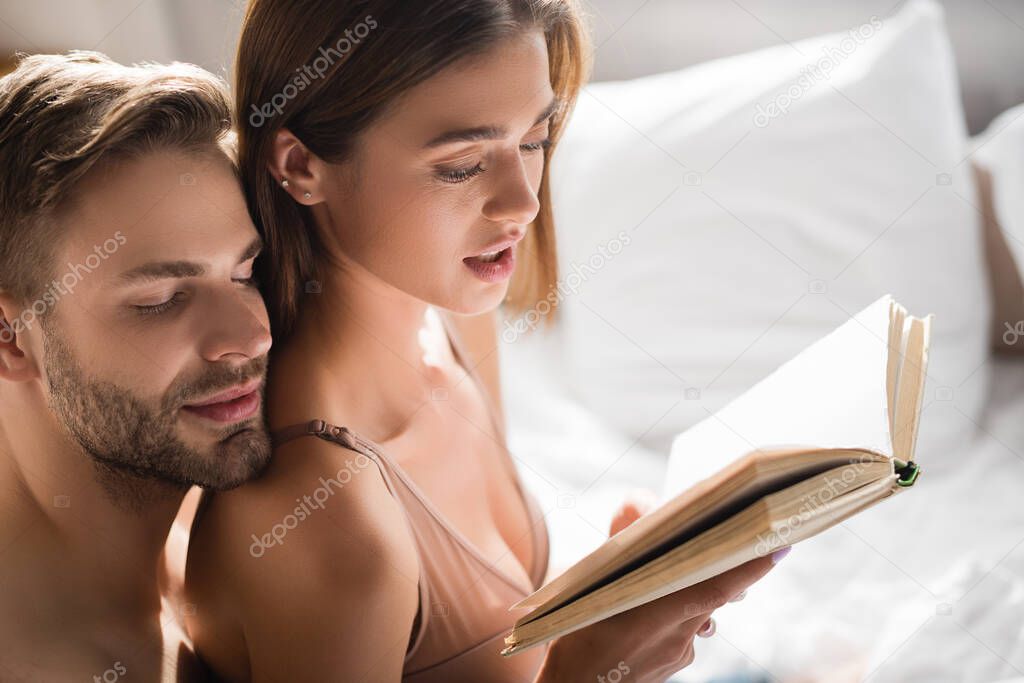 happy man leaning on shoulder of sexy girlfriend reading book in bedroom