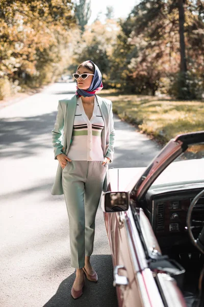 Elegant woman in sunglasses with hands in pockets looking away near car on blurred foreground on road