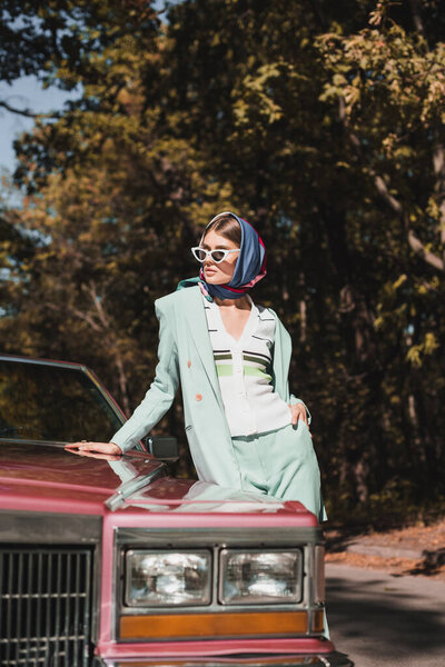 Stylish woman with hand in pocket standing beside vintage car on blurred foreground