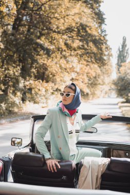 Stylish woman in headscarf looking away on driver seat of roofless car  clipart