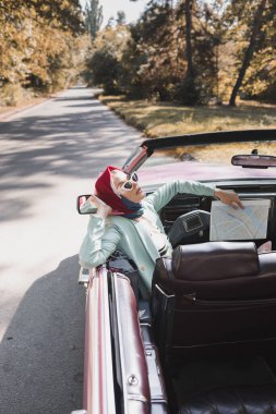 Fashionable woman with map sitting in roofless car on road during trip  clipart