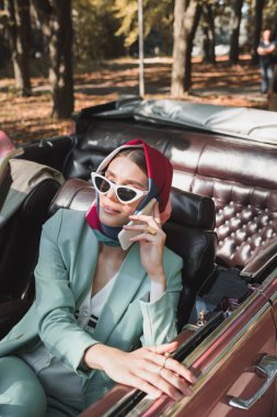 Smiling elegant woman talking on smartphone in roofless car during trip  clipart