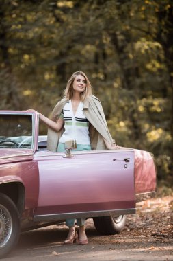 young woman looking away while standing near cabriolet on roadside clipart