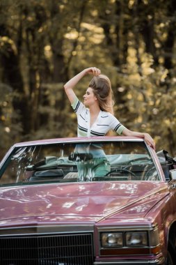 stylish woman touching hair and looking away while standing in vintage cabriolet clipart