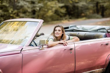 cheerful young woman looking away while sitting in convertible car clipart