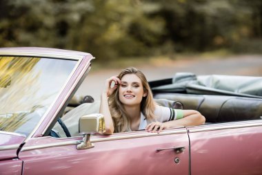 happy young woman holding hand near face while sitting in cabriolet and looking at camera clipart