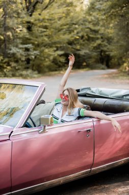 Güneş gözlüklü, heyecanlı genç bir kadın. Retro Cabriolet 'te oturuyor. Elleri havada.