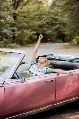Neşeli bir kadın Cabriolet 'te otururken kafası kalkık, gözleri kapalı ve eli havada oturuyor.