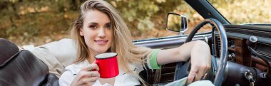 Üstü açık bir arabada elinde kahve fincanı tutarken kameraya gülümseyen mutlu genç bir kadın.