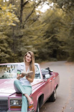 young smiling woman sitting on hood of vintage cabriolet and reading book clipart