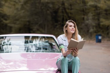 happy woman looking away while sitting on hood of cabriolet and looking away clipart