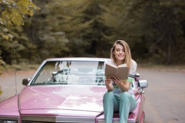 happy young woman reading book while sitting on hood of convertible car on blurred background clipart
