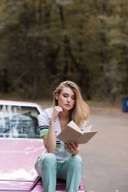 Cabriolet 'in kaputunda oturan ve bulanık arka plan üzerine kitap okuyan düşünceli bir kadın.