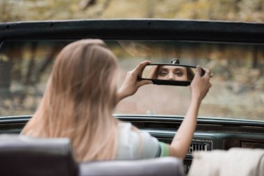 back view of young woman adjusting rearview mirror in cabriolet on blurred foreground clipart