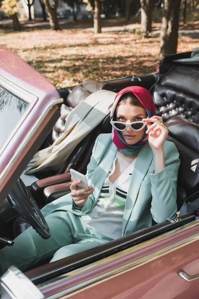 戴着太阳镜 手持智能手机 坐在复古车上看着相机的女人前景暗淡 — 图库照片