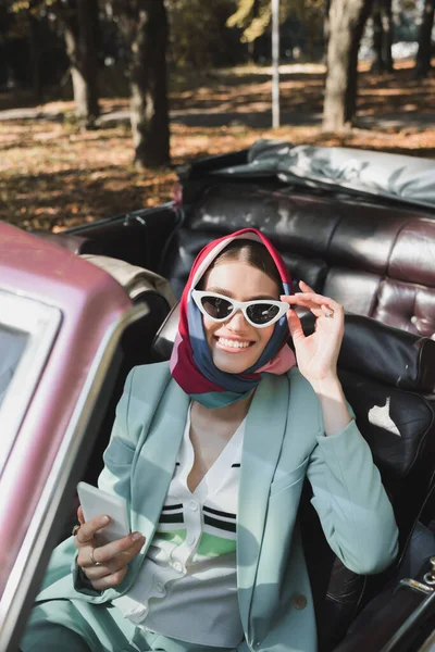 Mujer Sonriente Tocando Gafas Sol Mientras Sostiene Teléfono Inteligente Coche — Foto de Stock