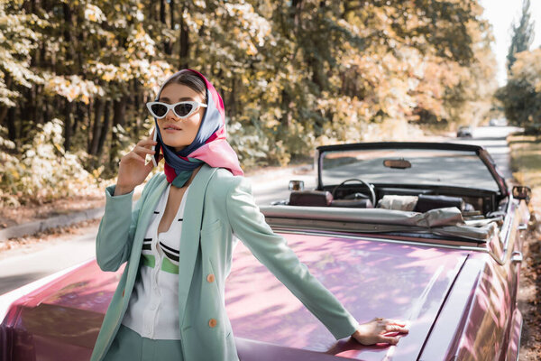 Elegant woman talking on smartphone near retro car on road during trip 