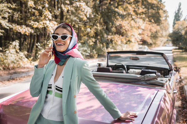 Smiling woman in sunglasses talking on smartphone near cabriolet car on road 