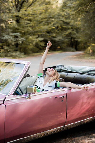 joyful woman relaxing in cabriolet while sitting with raised head, closed eyes and hand in air
