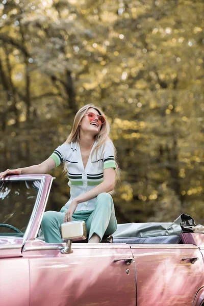 Podekscytowana Kobieta Okularach Przeciwsłonecznych Odwracając Wzrok Podczas Pozowania Samochodzie Lesie — Zdjęcie stockowe