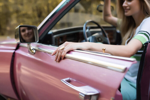 cropped view of woman opening door of cabriolet while looking in side view mirror on blurred background