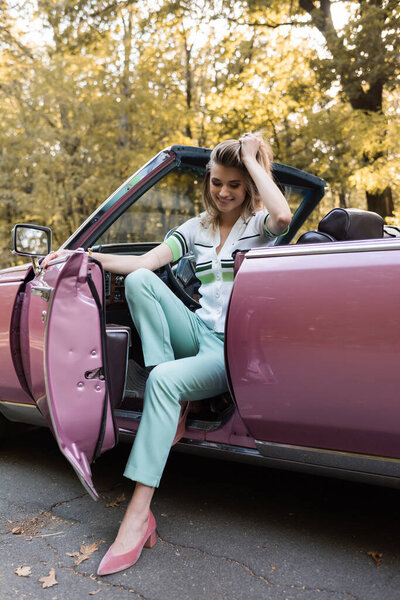 happy stylish woman touching hair while stepping out cabriolet