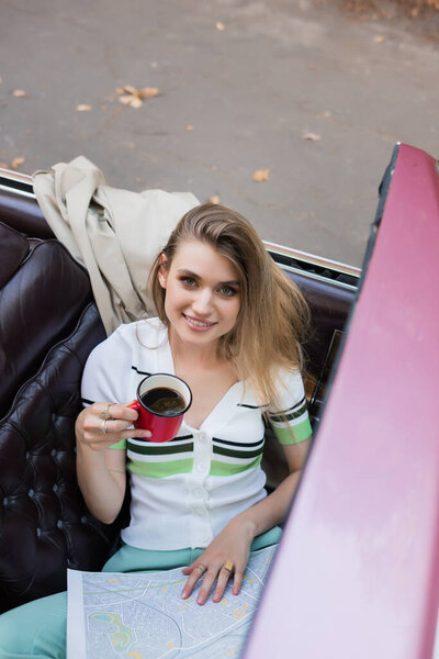 overhead view of young woman smiling at camera while holding cup of coffee and road atlas in cabriolet