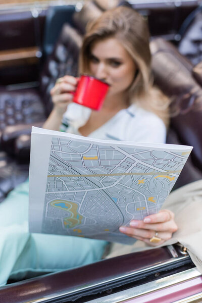 young woman looking at road atlas and drinking coffee while sitting in cabriolet on blurred background