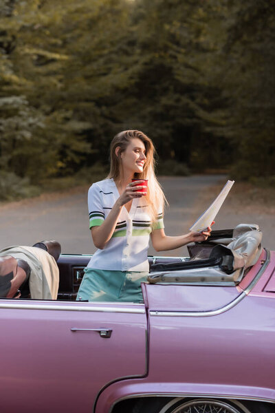 happy woman holding map and cup of coffee while standing in convertible car