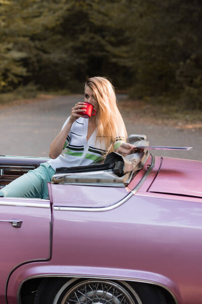 stylish woman looking away while drinking coffee and holding road map in vintage cabriolet